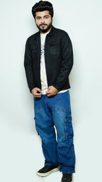 Man wearing a black jacket, white shirt, and blue jeans against a light gray background